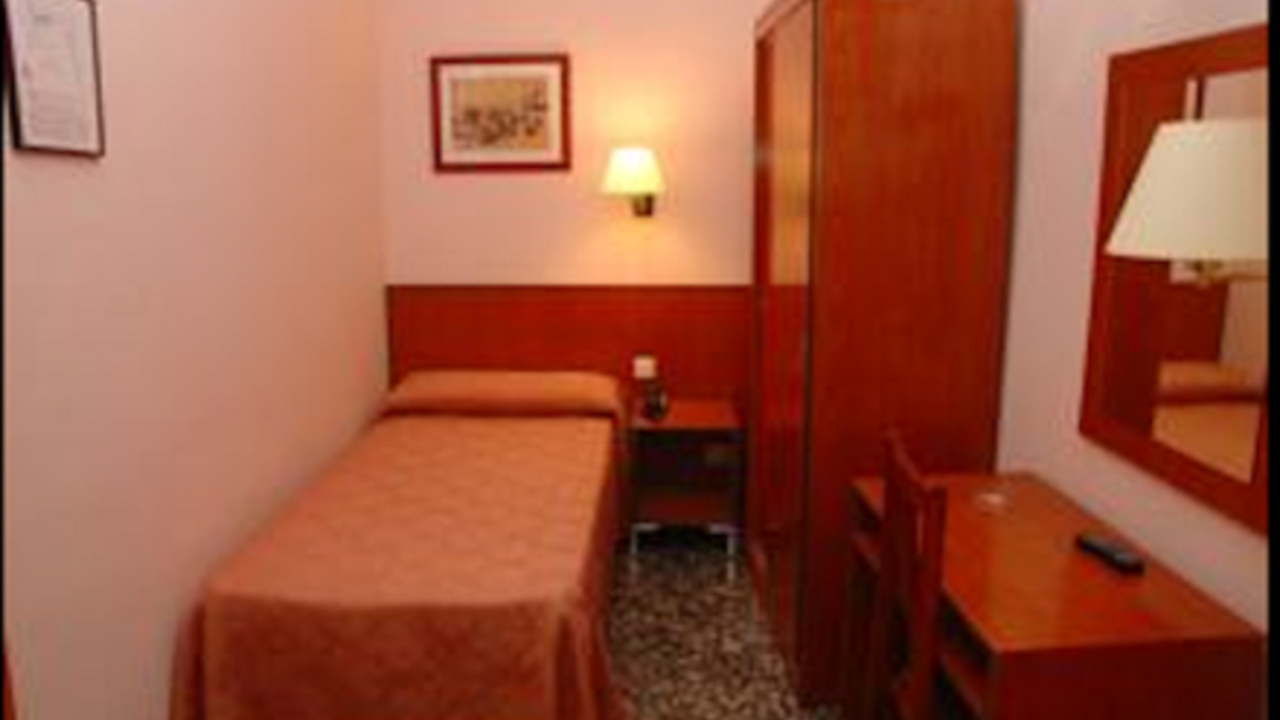 Hotel room with single bed, desk, wardrobe, and nightstand. Pink walls, brown furniture.