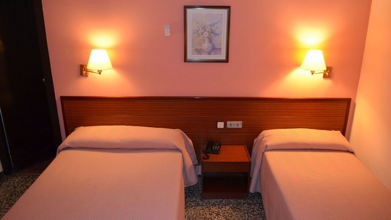 Two twin beds in a hotel room with a nightstand, lamps, and artwork on a pink wall.