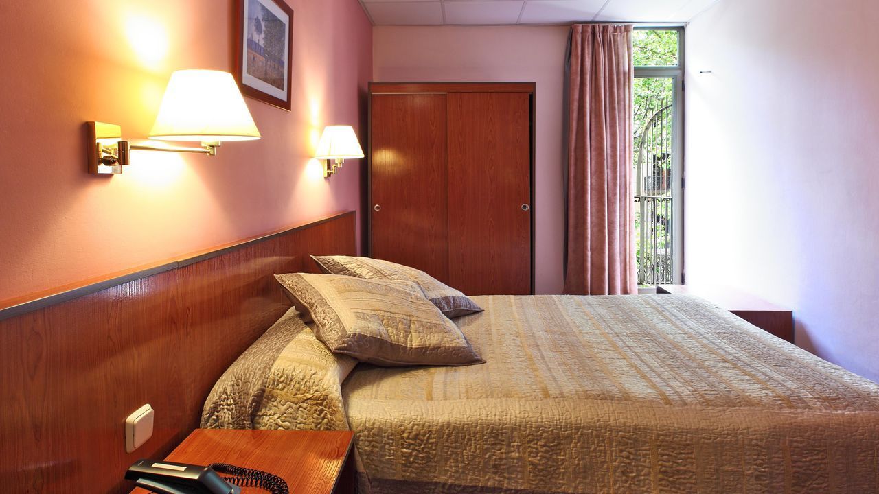 Hotel bedroom with a bed, wardrobe, and window. Pink walls, brown headboard, and two wall lamps.