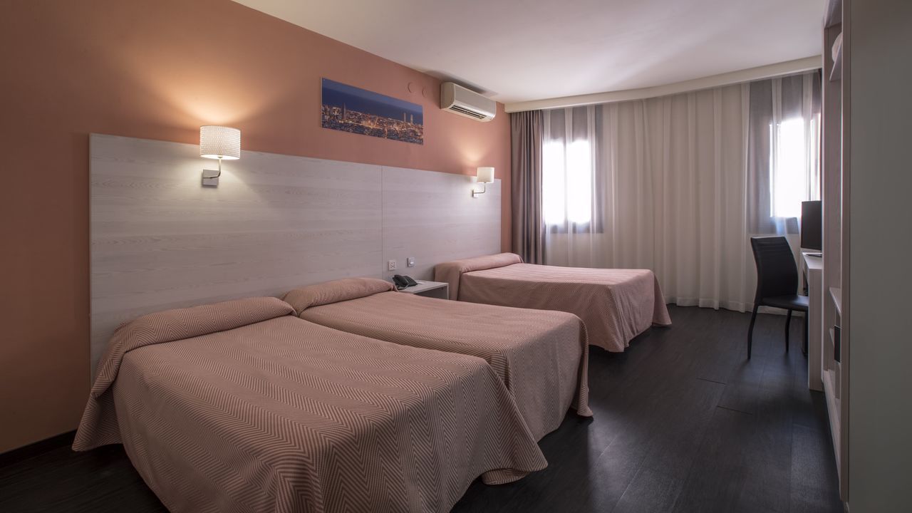 Hotel room with three beds, pale pink bedding, peach walls, and dark floors.