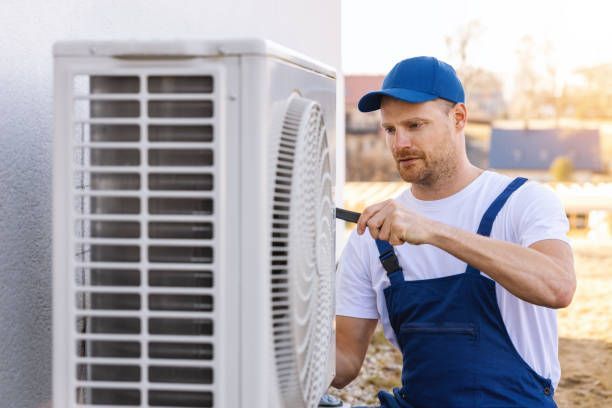 HVAC technician in blue overalls and cap working on an outdoor air conditioning unit.