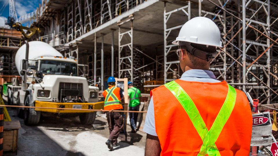 Construction worker in orange vest and white hardhat, observing a concrete mixer truck on a construction site.