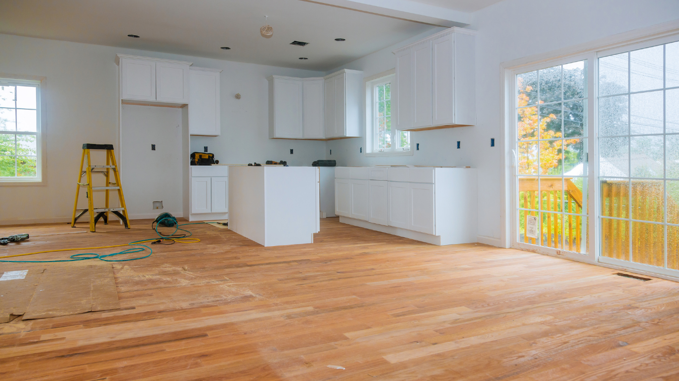 Kitchen renovation in progress, white cabinets, island, wood floor, ladder, tools, and a sliding glass door.