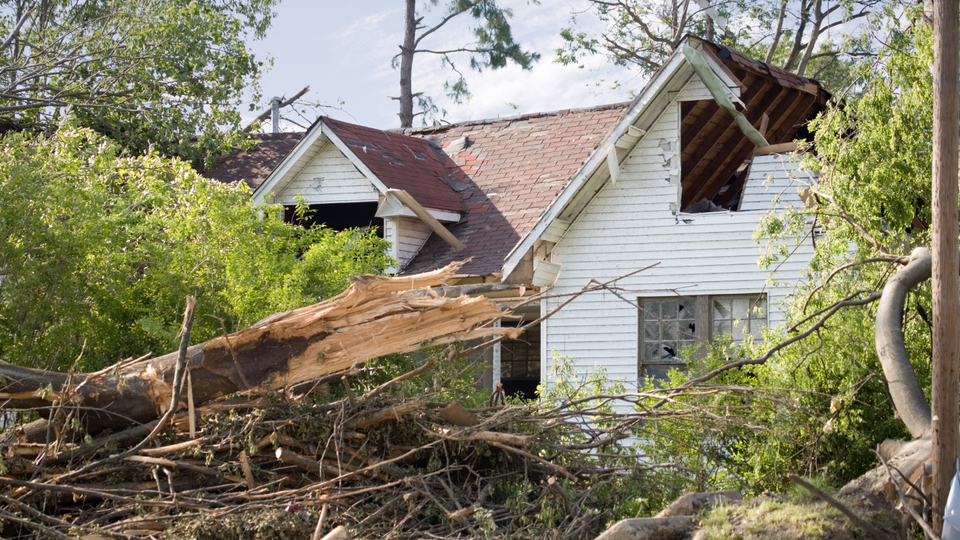 House heavily damaged by a storm, surrounded by debris and trees.