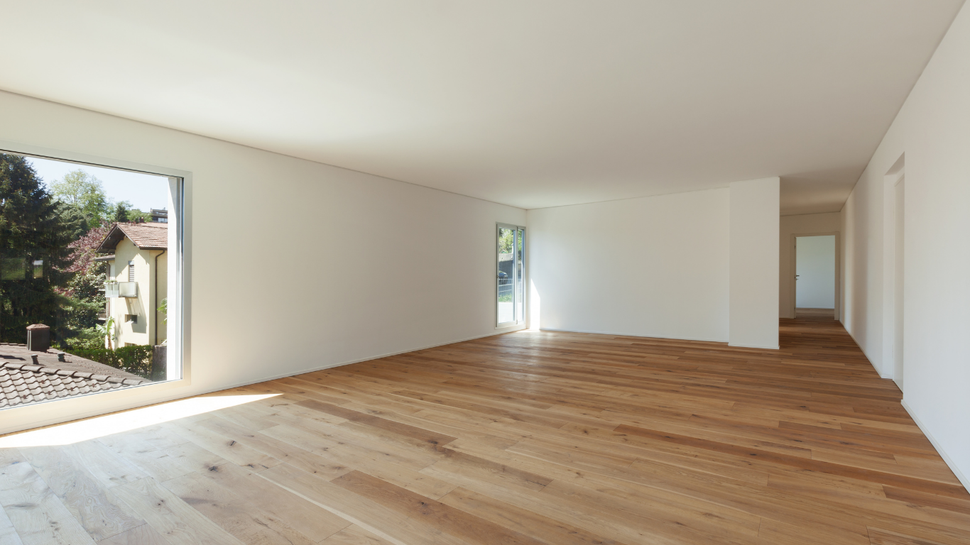 Empty room with wooden floors, white walls, and a large window overlooking trees.