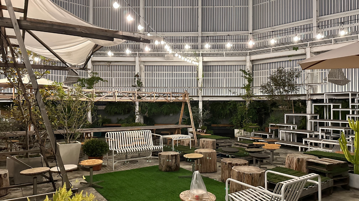 Outdoor cafe with seating on grass, surrounded by greenery and a striped building facade.