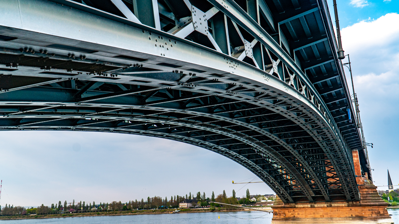 Underside view of a large, arched metal bridge over water; gray and blue tones, blue sky background.