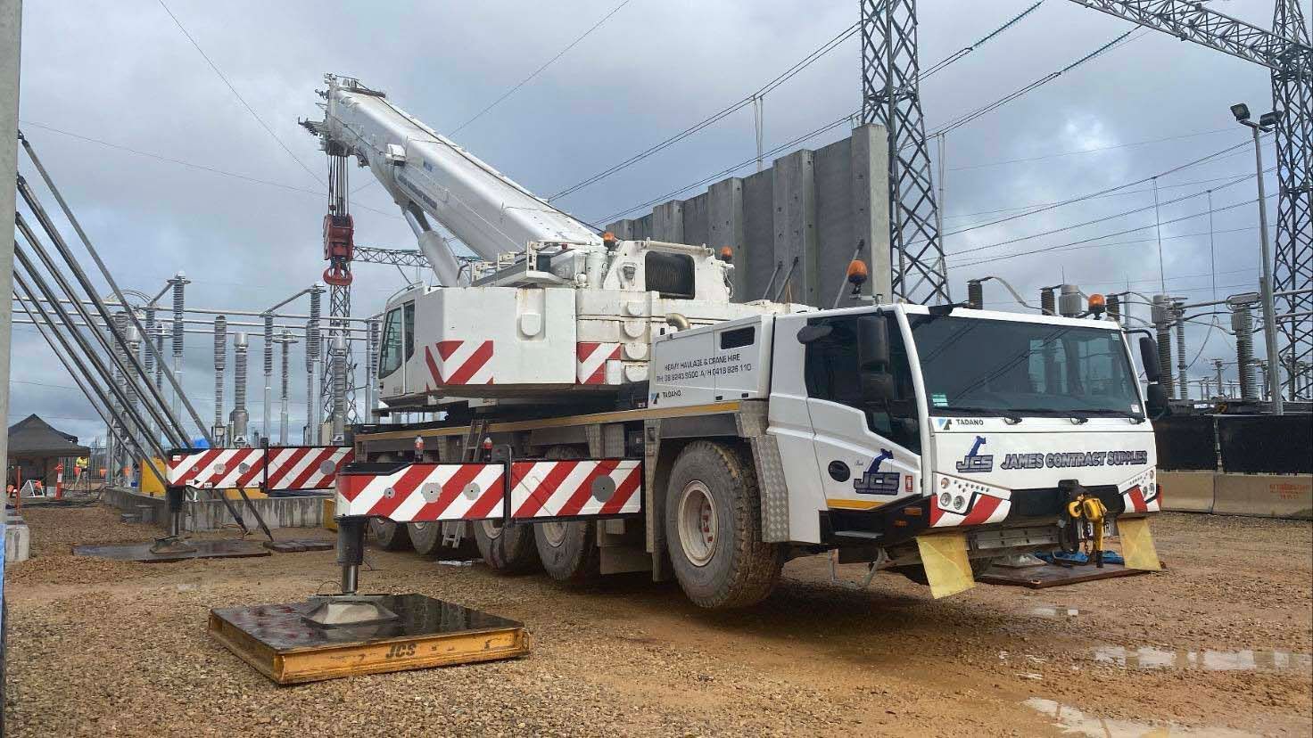 A large white truck with a crane attached to it is parked in front of a power station.