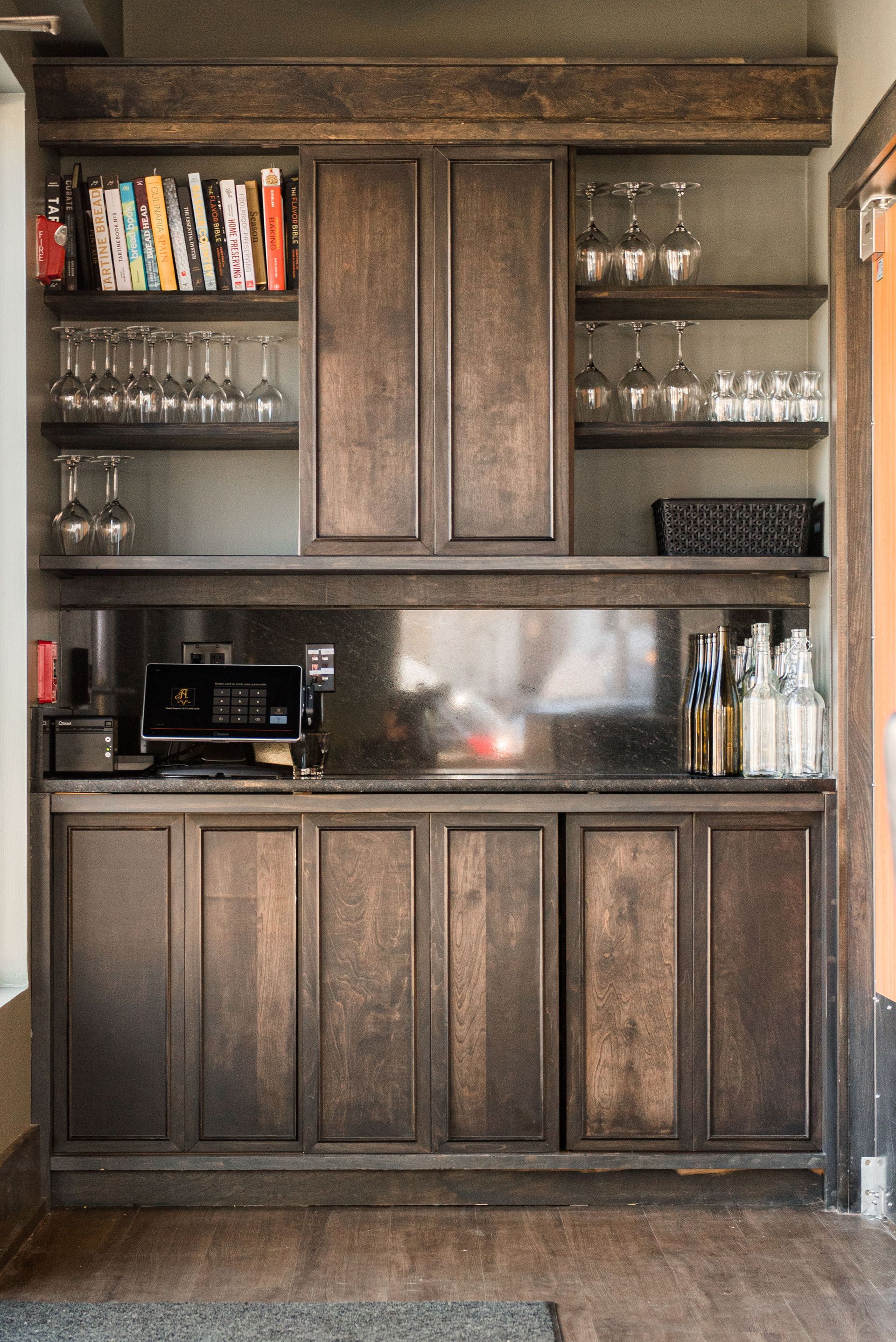 A kitchen with wooden cabinets and shelves filled with wine glasses and books.
