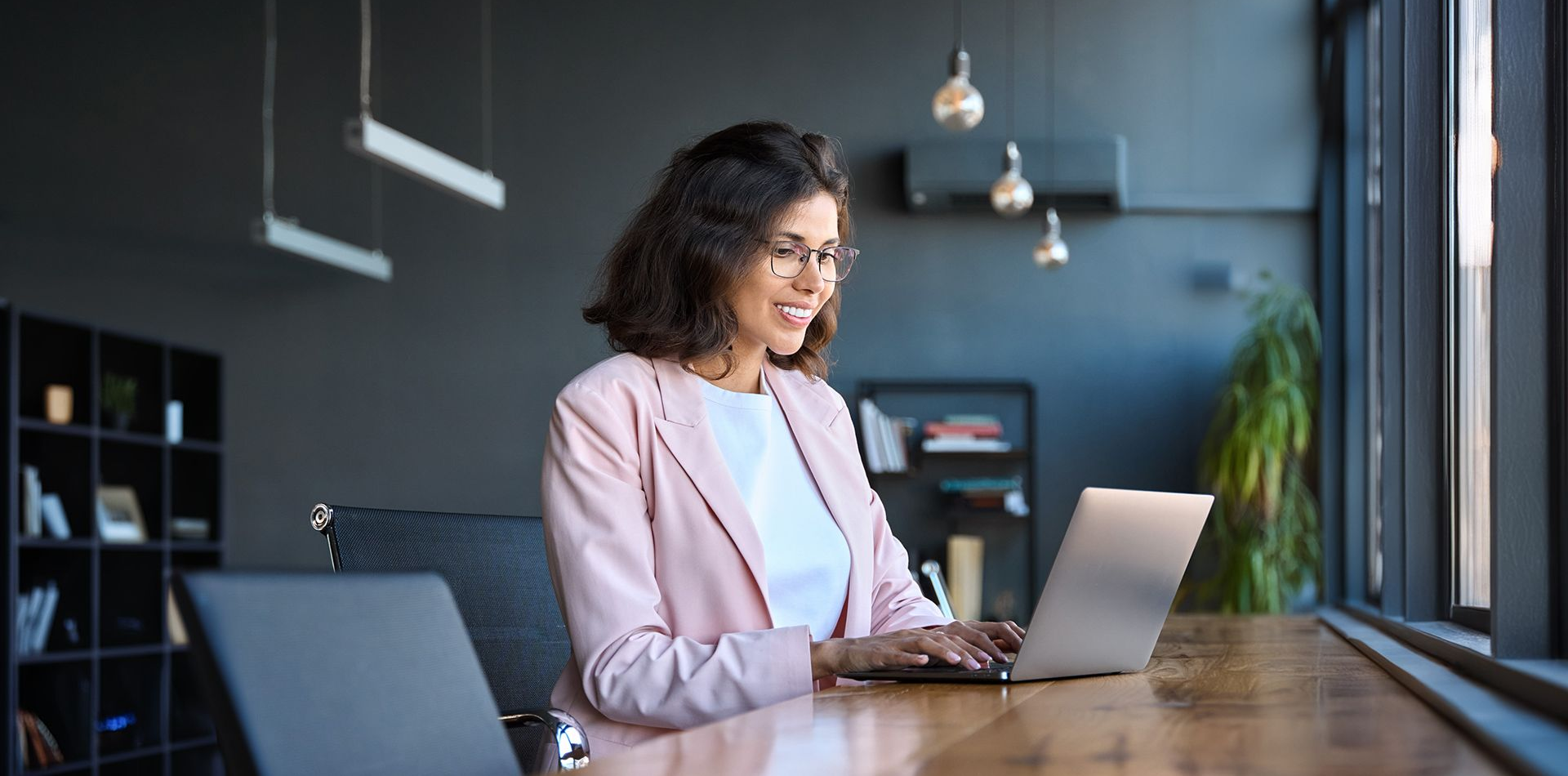 Woman working on a laptop at a desk in a modern office, with hanging lights and a plant in the background