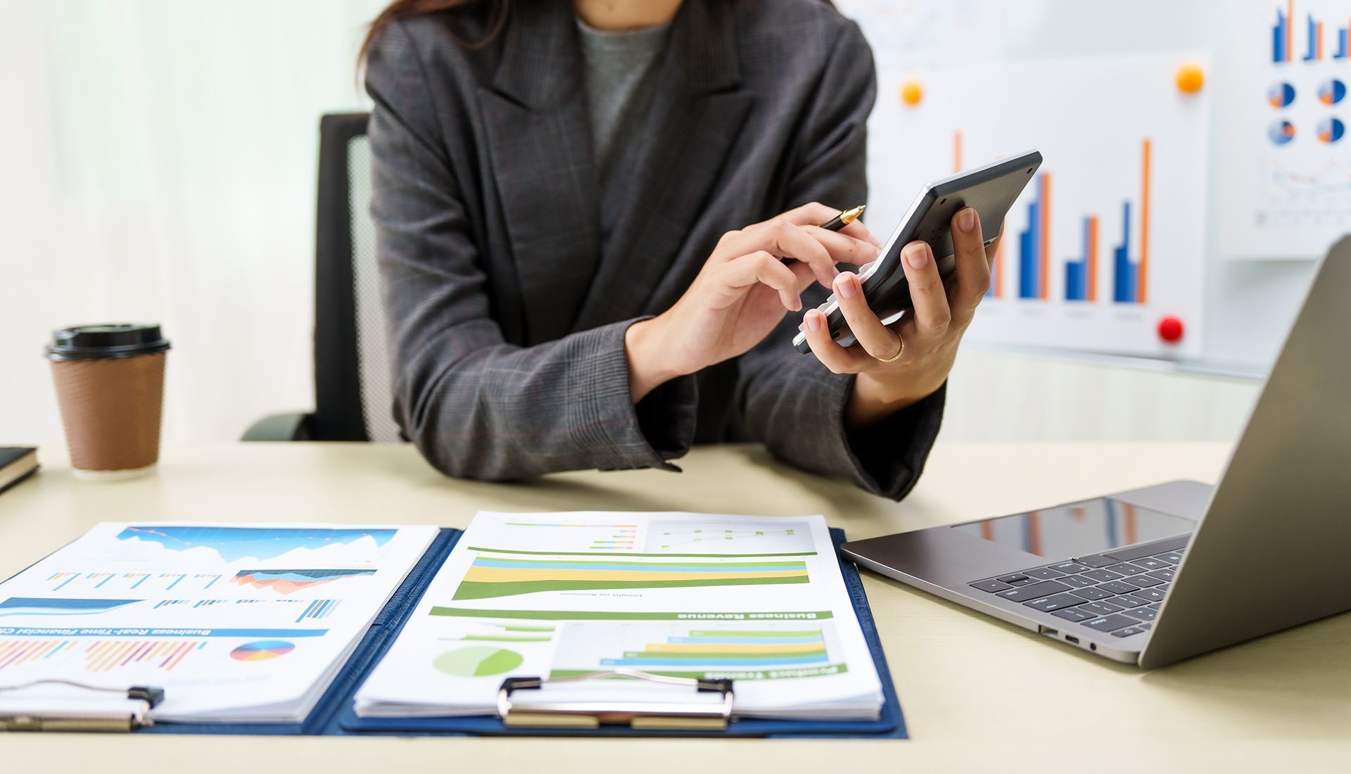 Woman in a blazer using a calculator, looking at charts and a laptop in an office setting.