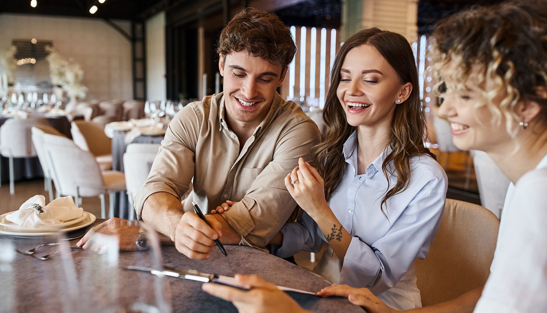 Three people smiling around a restaurant table; one writing, two observing.
