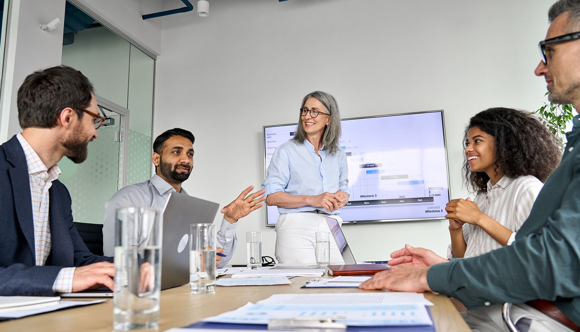 Business team meeting around a table, presentation on screen.