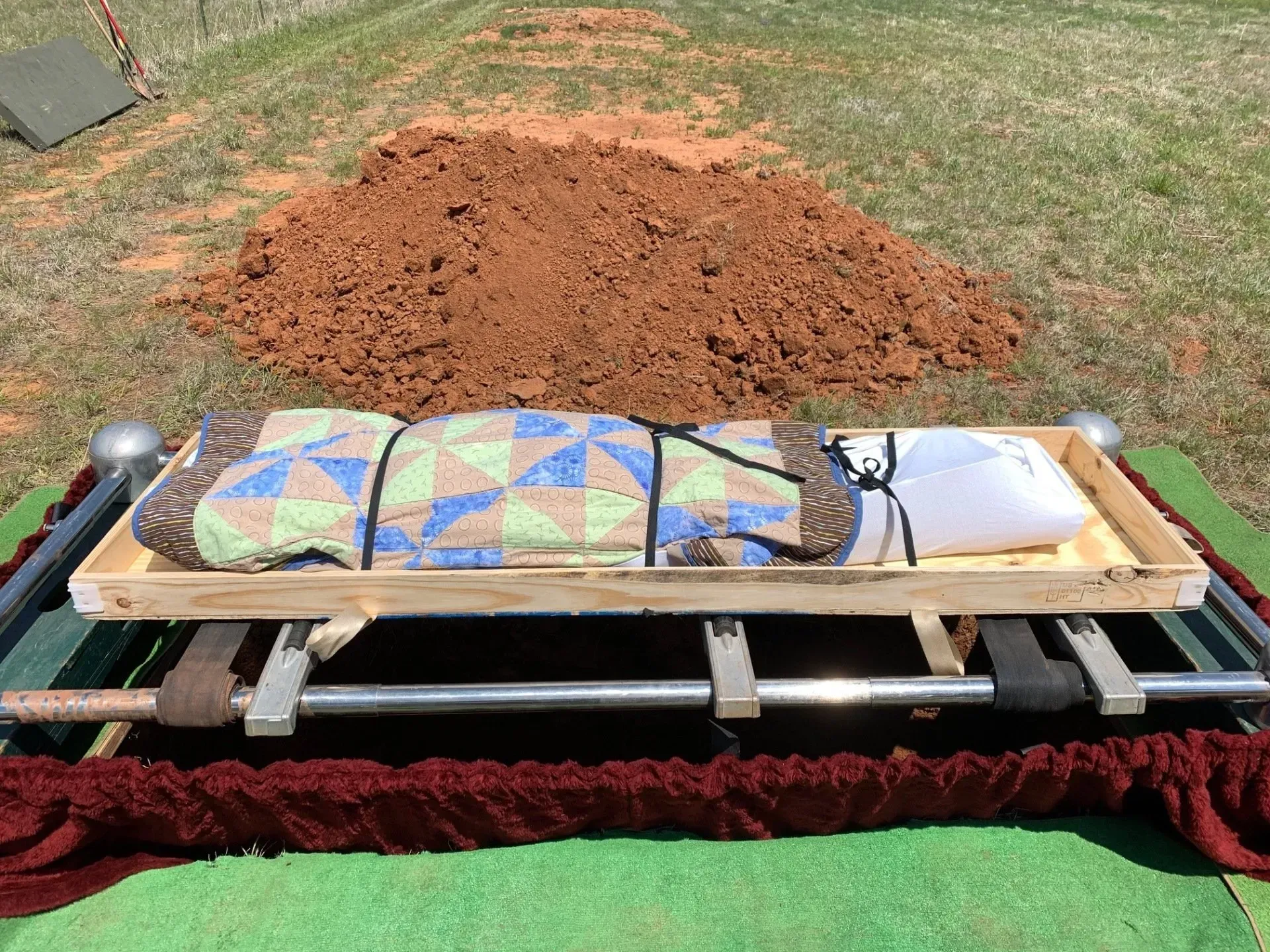 Two colorful quilts laid on a wooden bench in front of a large pile of dirt outdoors.