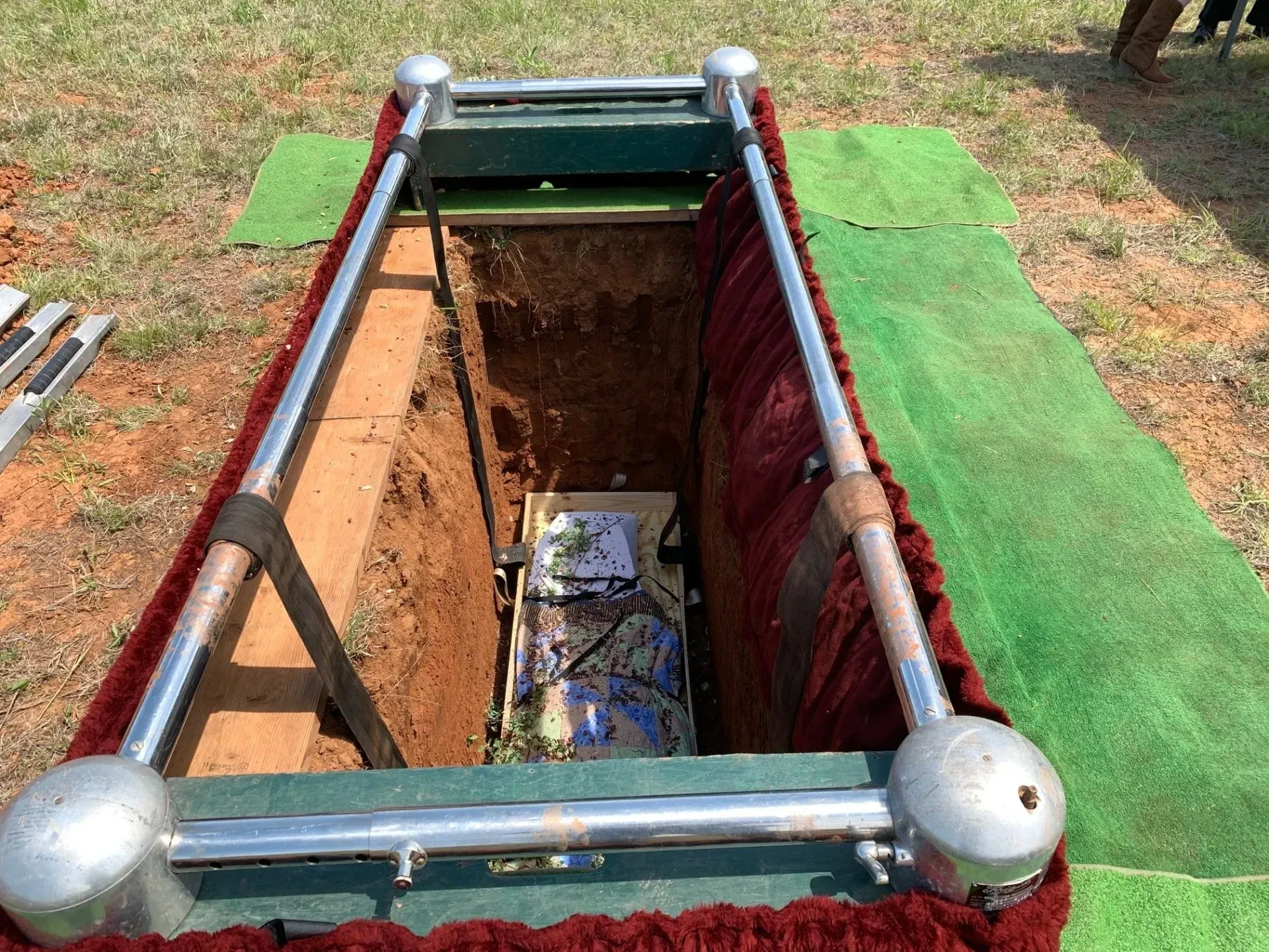 Open grave with coffin and red lining, surrounded by metal rails and green turf decorations