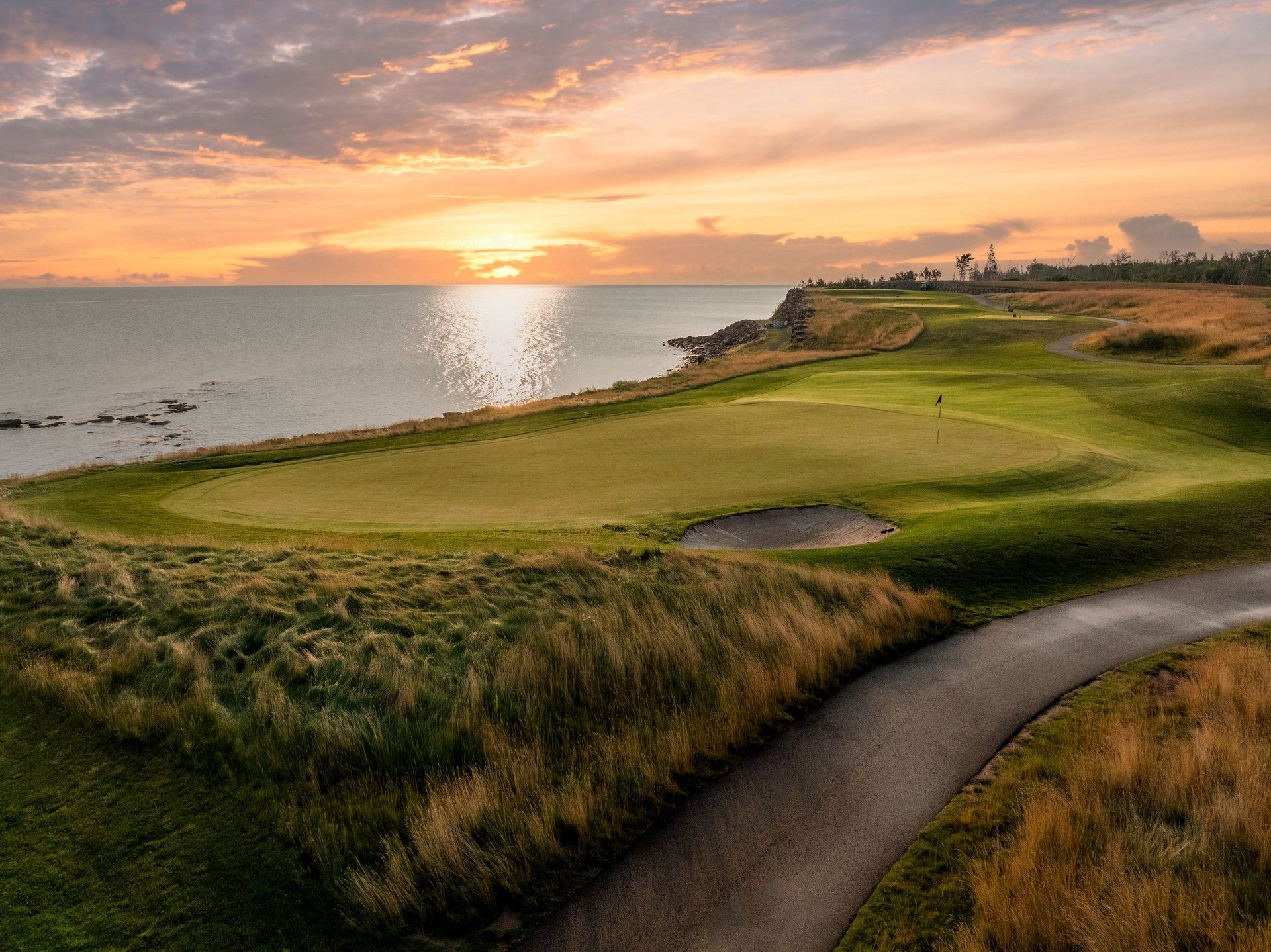 Golf course by ocean at sunset; green grass, sand trap, path, sun reflecting on water.