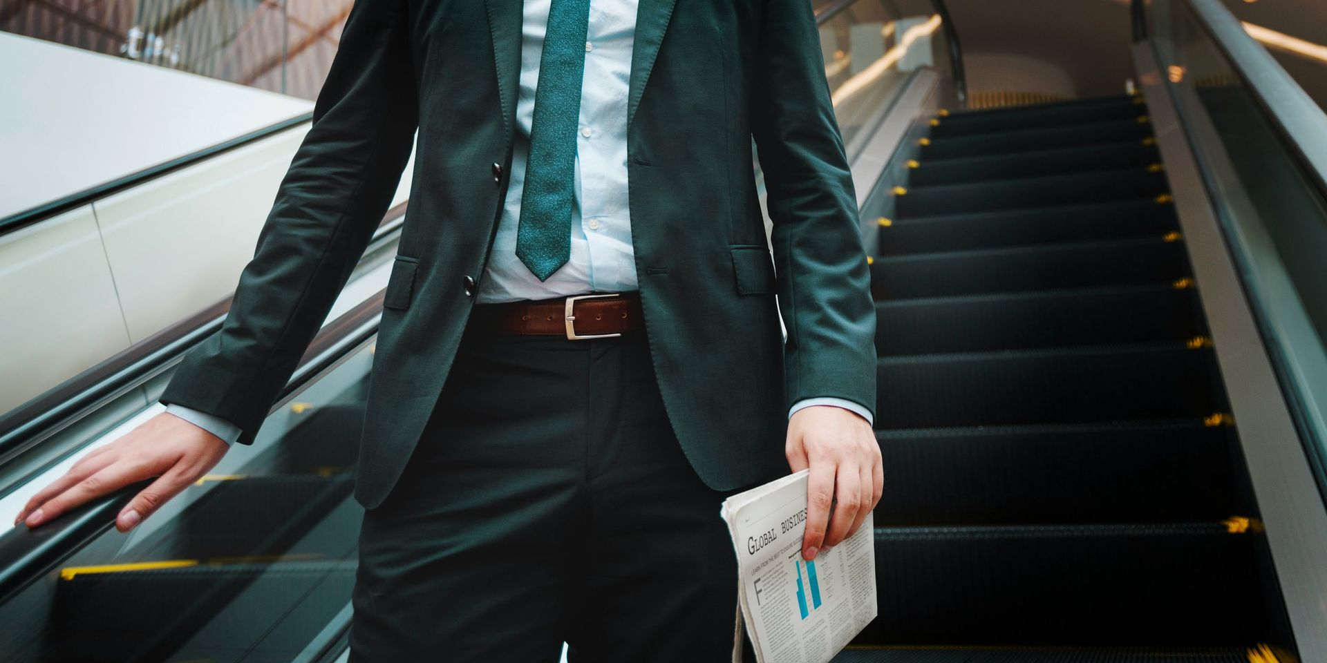 Person in suit riding up an escalator, holding a document.