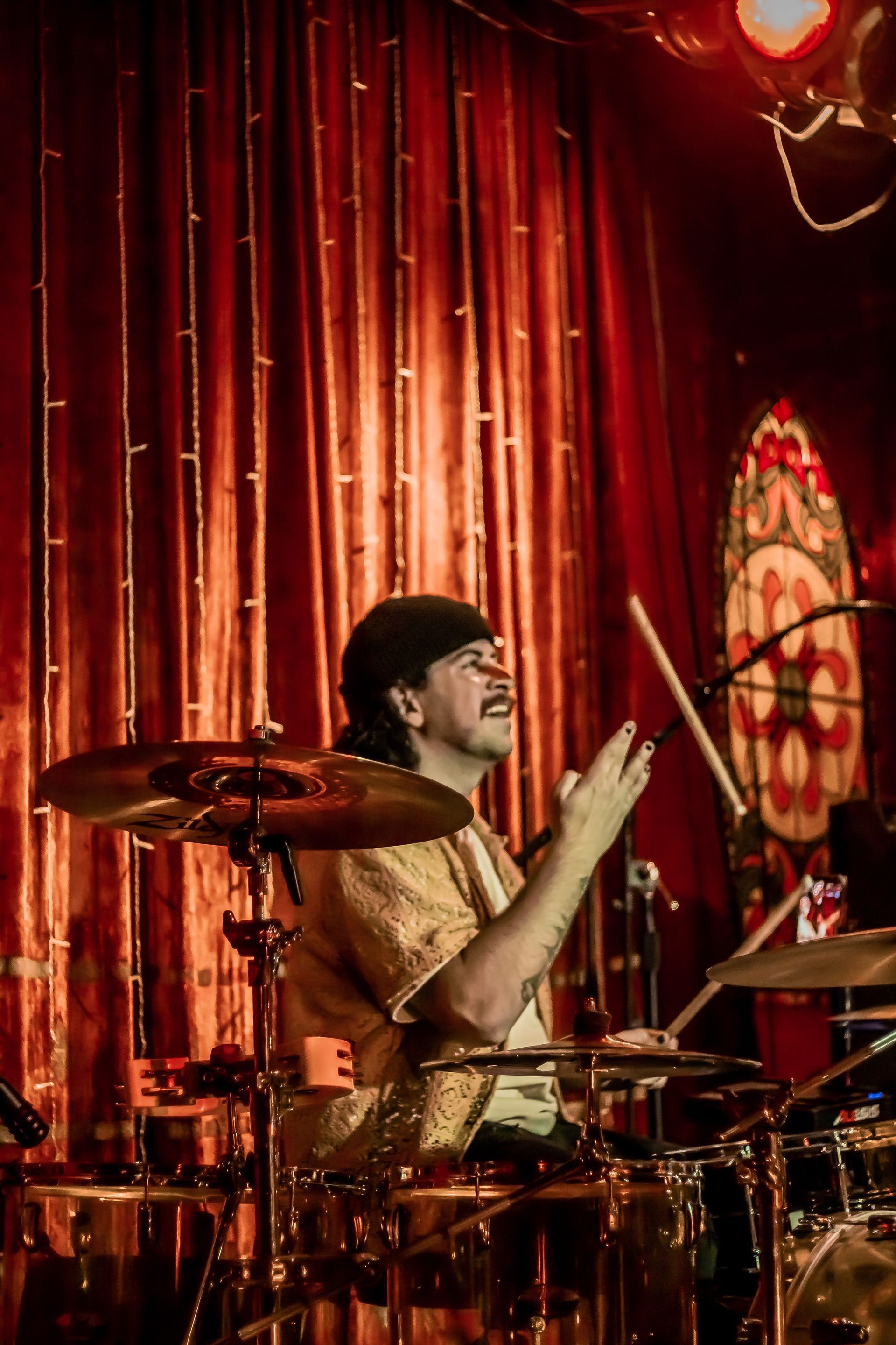 Mikey from 19 Miles Per Hour is playing drums on a stage in front of a red curtain.