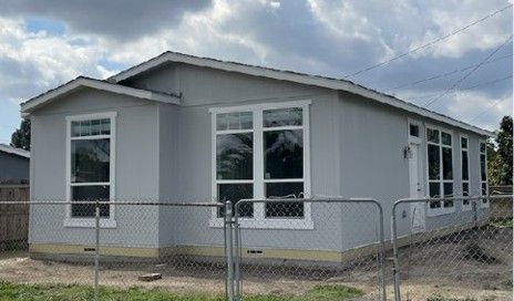 Light gray house with large windows, behind a chain-link fence, under a cloudy sky.