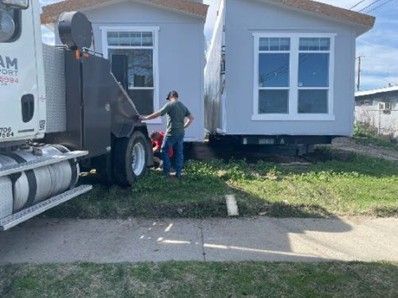A house being transported by a truck on a grassy area, with a person inspecting the connection.