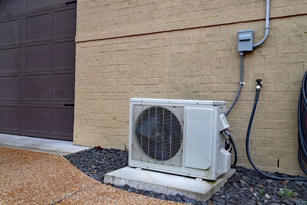 Outdoor HVAC condenser unit beside a tan brick house wall with utility boxes and pipes