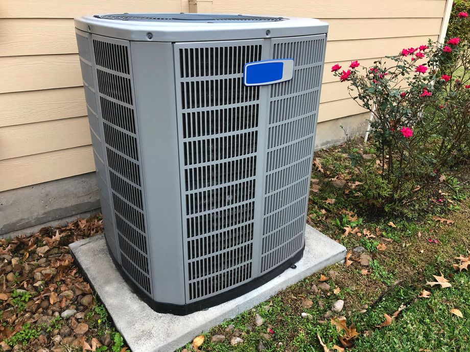 Outdoor air conditioner unit beside a beige house wall and pink flowers on a concrete pad