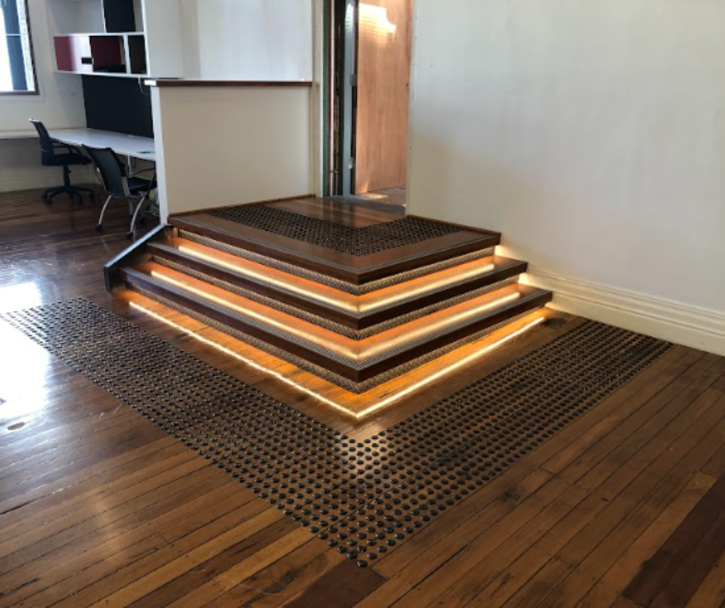 Illuminated tiered wooden platform with handrail on a woven rug in an office space