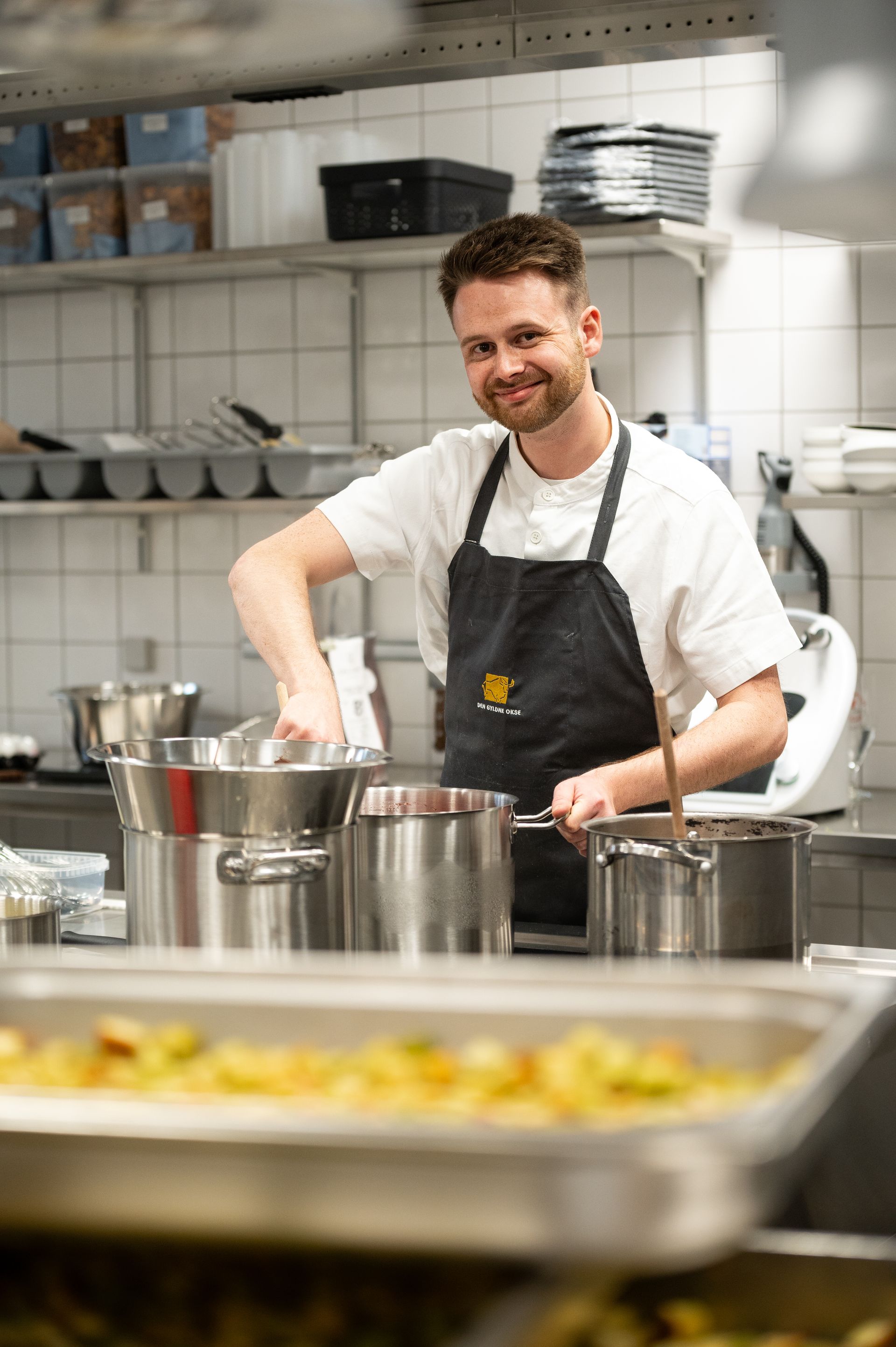 Kok i hvid skjorte og forklæde smiler, mens han rører i maden i et køkken.