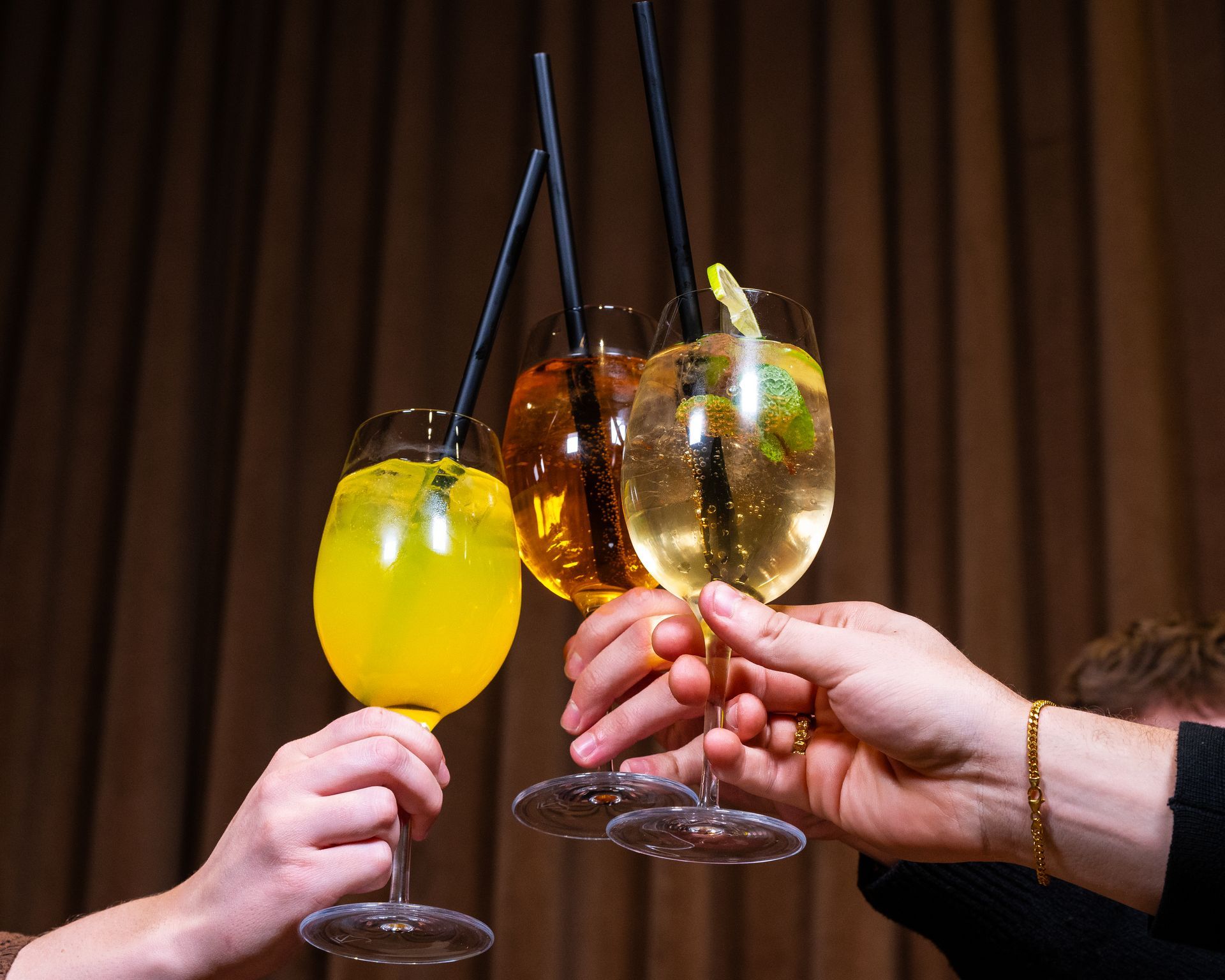 Hænder klirrer vinglas fyldt med cocktails, med sugerør. Brun gardinbaggrund.