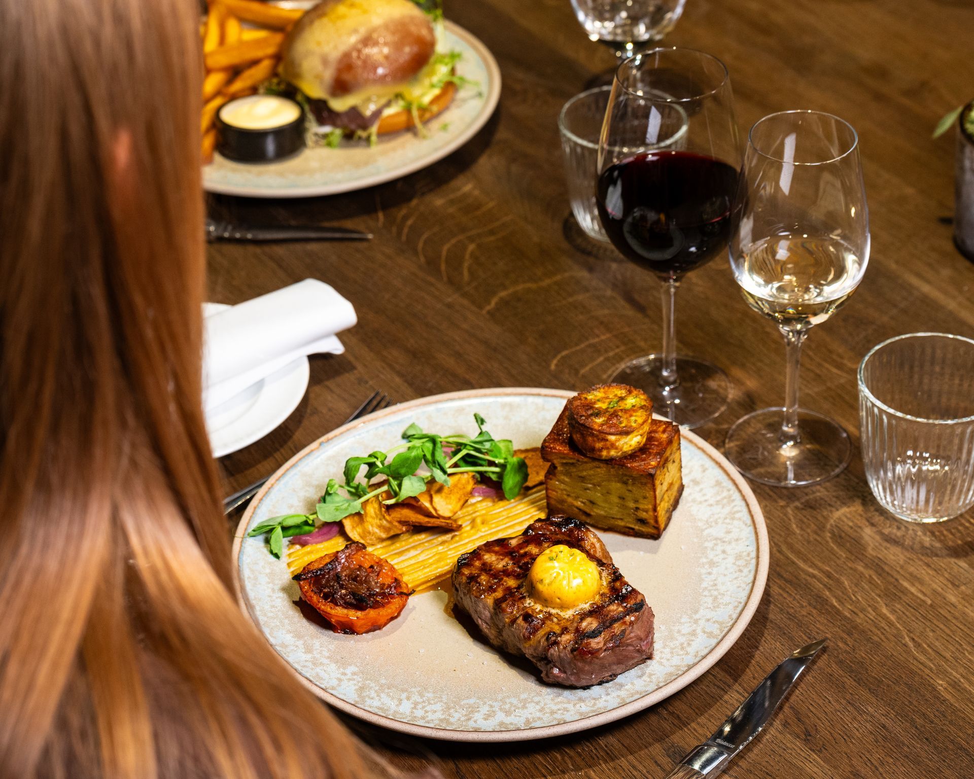 Person ved et restaurantbord med bøf, grøntsager, vinglas og en burger i baggrunden.