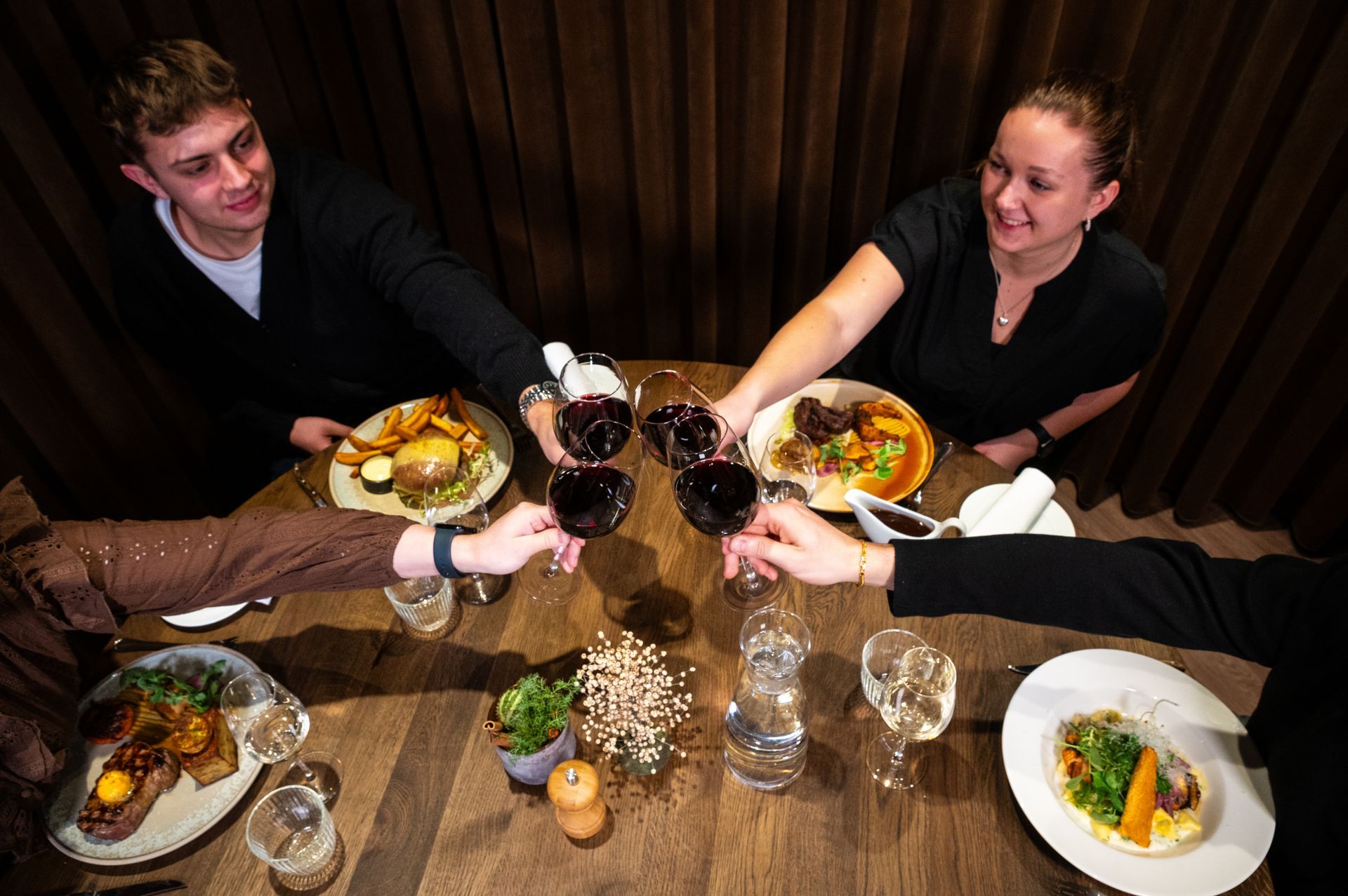 Folk skåler med vinglas omkring et træbord fyldt med mad. Mørke omgivelser.