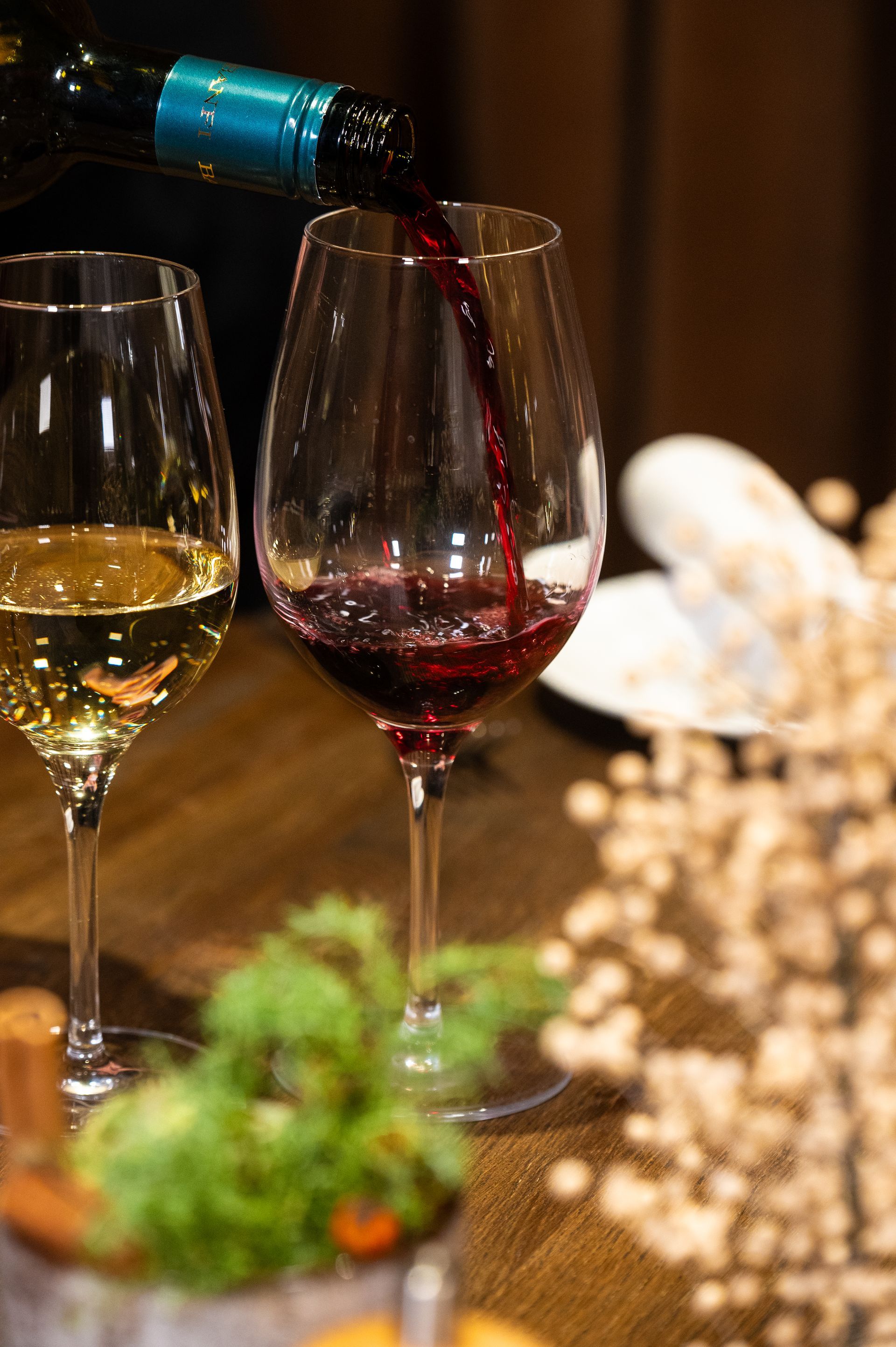 Rødvin hældes i et glas; et hvidvinsglas står ved siden af ​​det på en træoverflade.