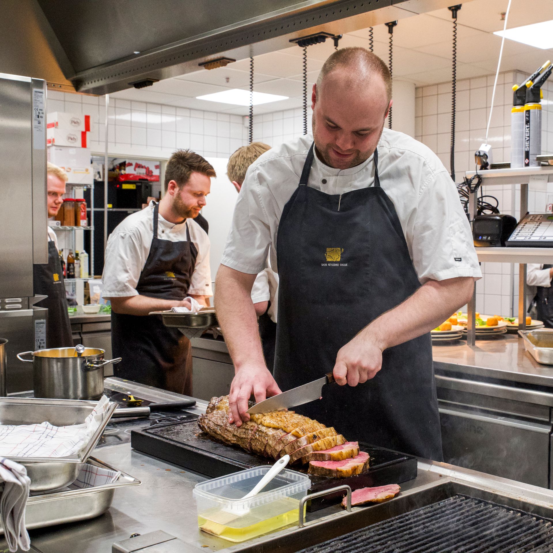 Kok skærer stegt kød i et travlt køkken, andre kokke arbejder i baggrunden.
