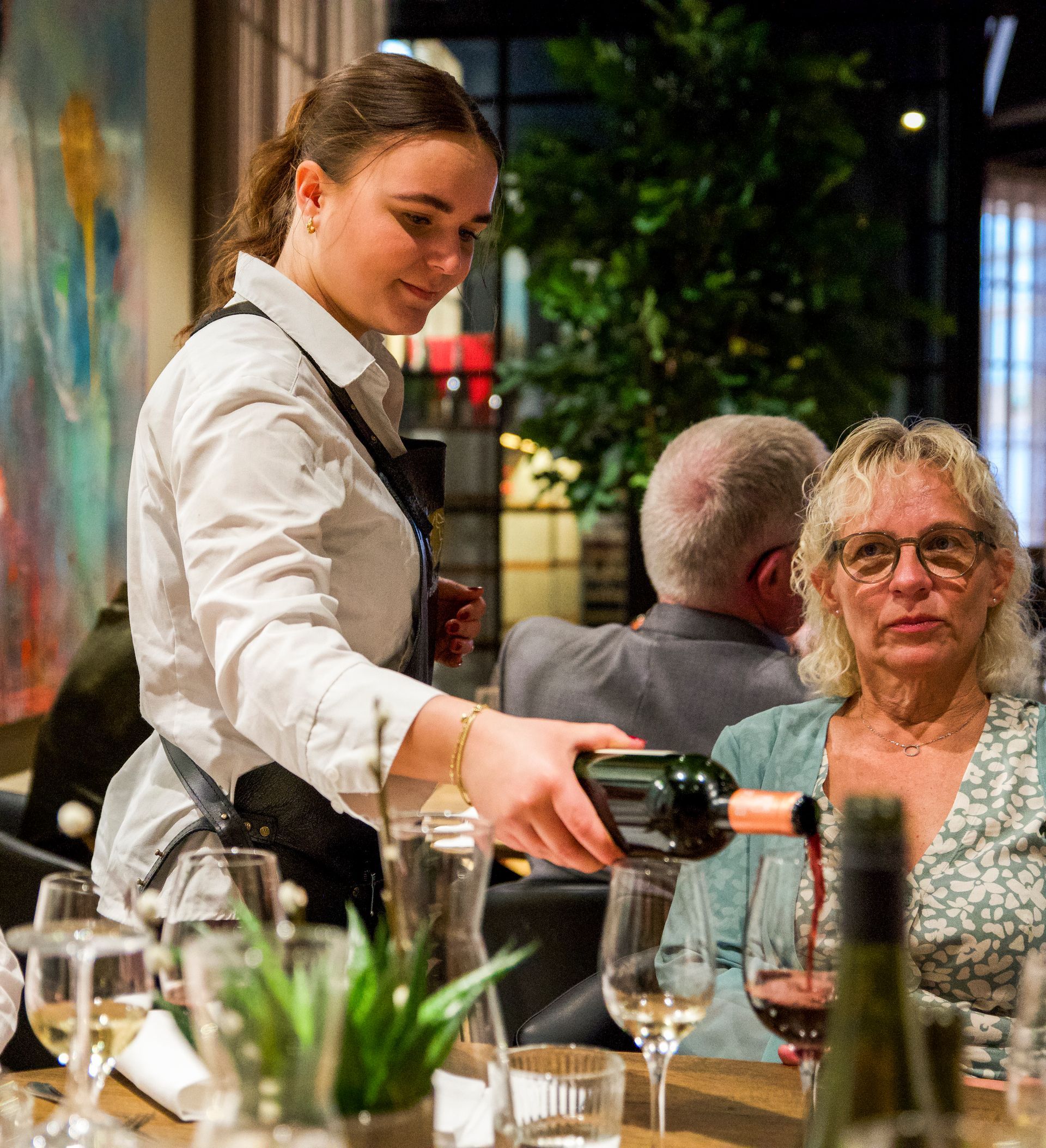 Servitrice hælder rødvin op til en kvinde ved et restaurantbord.
