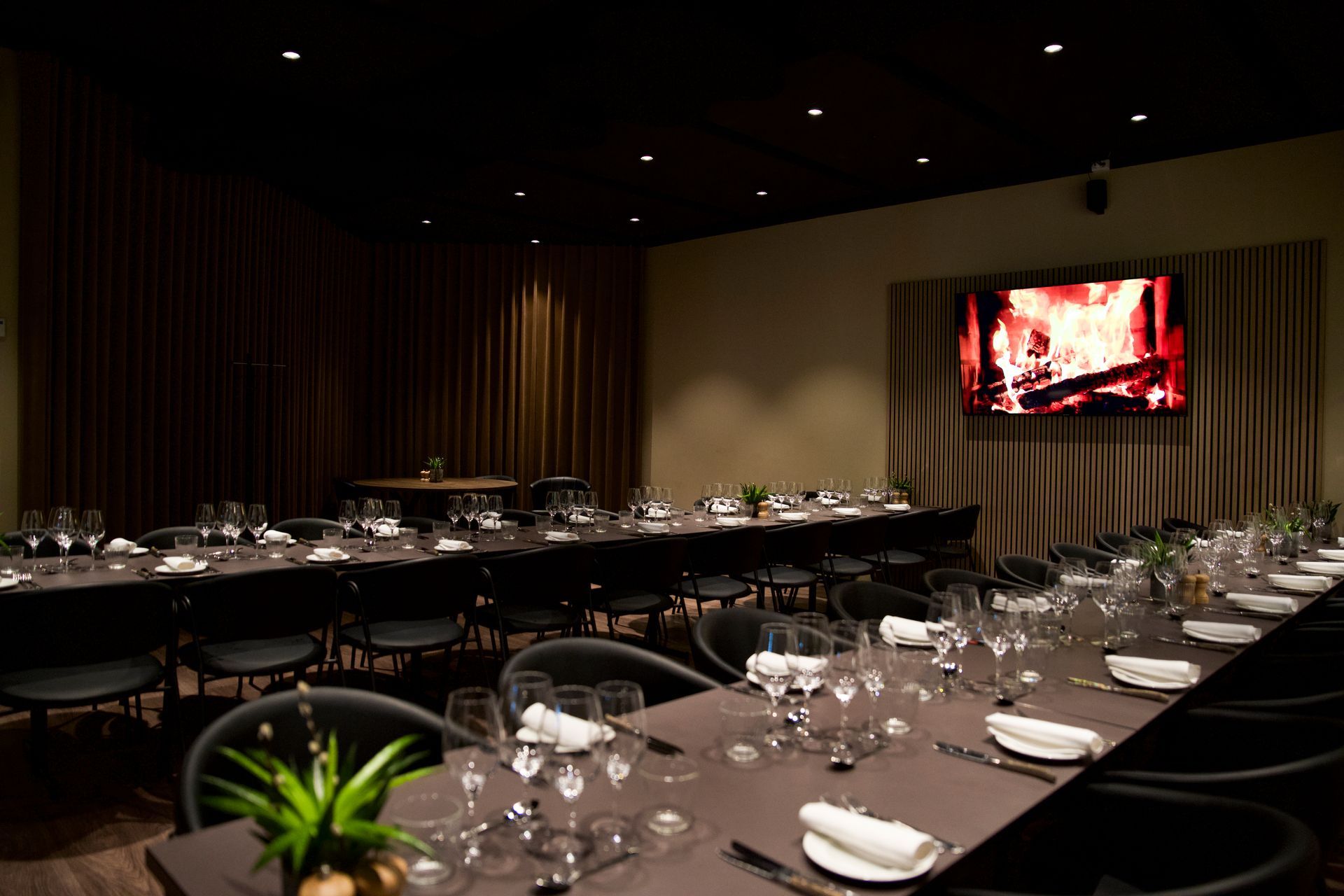 Formel spisestue til et stort arrangement; lange borde med kuverter, mørke stole, pejseudstilling på væggen.