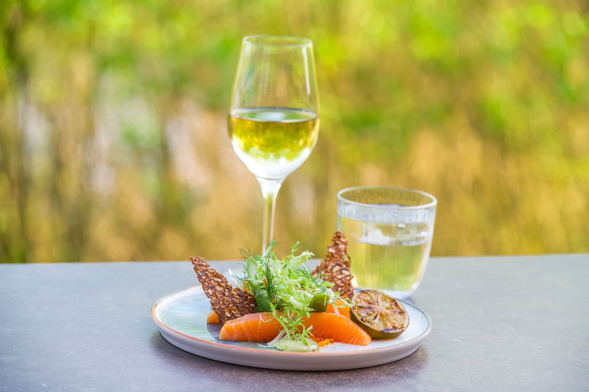 Tallerken med gulerødder, grønne krydderurter og chips, med vinglas og vandglas udendørs.