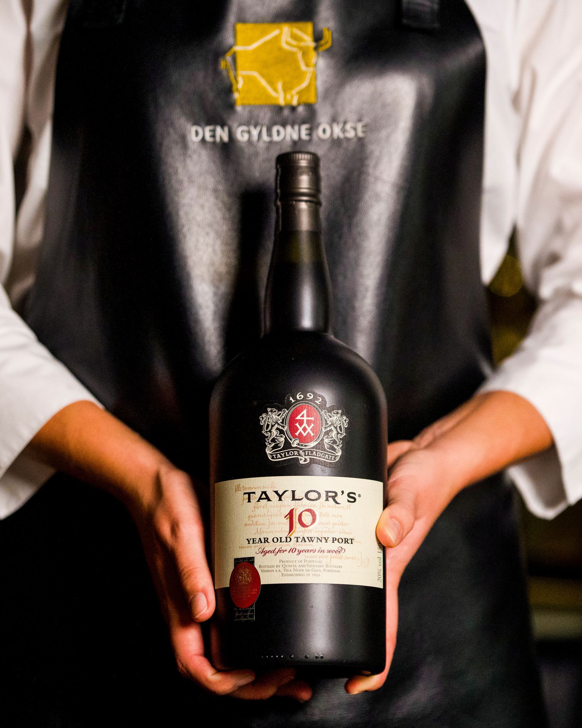 Person, der holder en Taylor's 10 Year Tawny Port-flaske foran et sort forklæde med et gyldent logo.