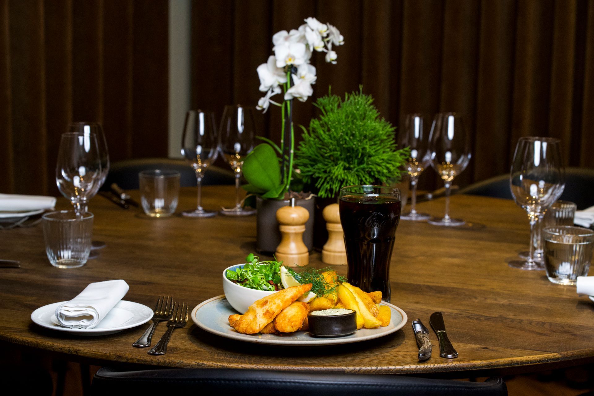 Fish and chips-måltid på et træbord, dækket til flere spisende gæster med vinglas og blomsterarrangement.