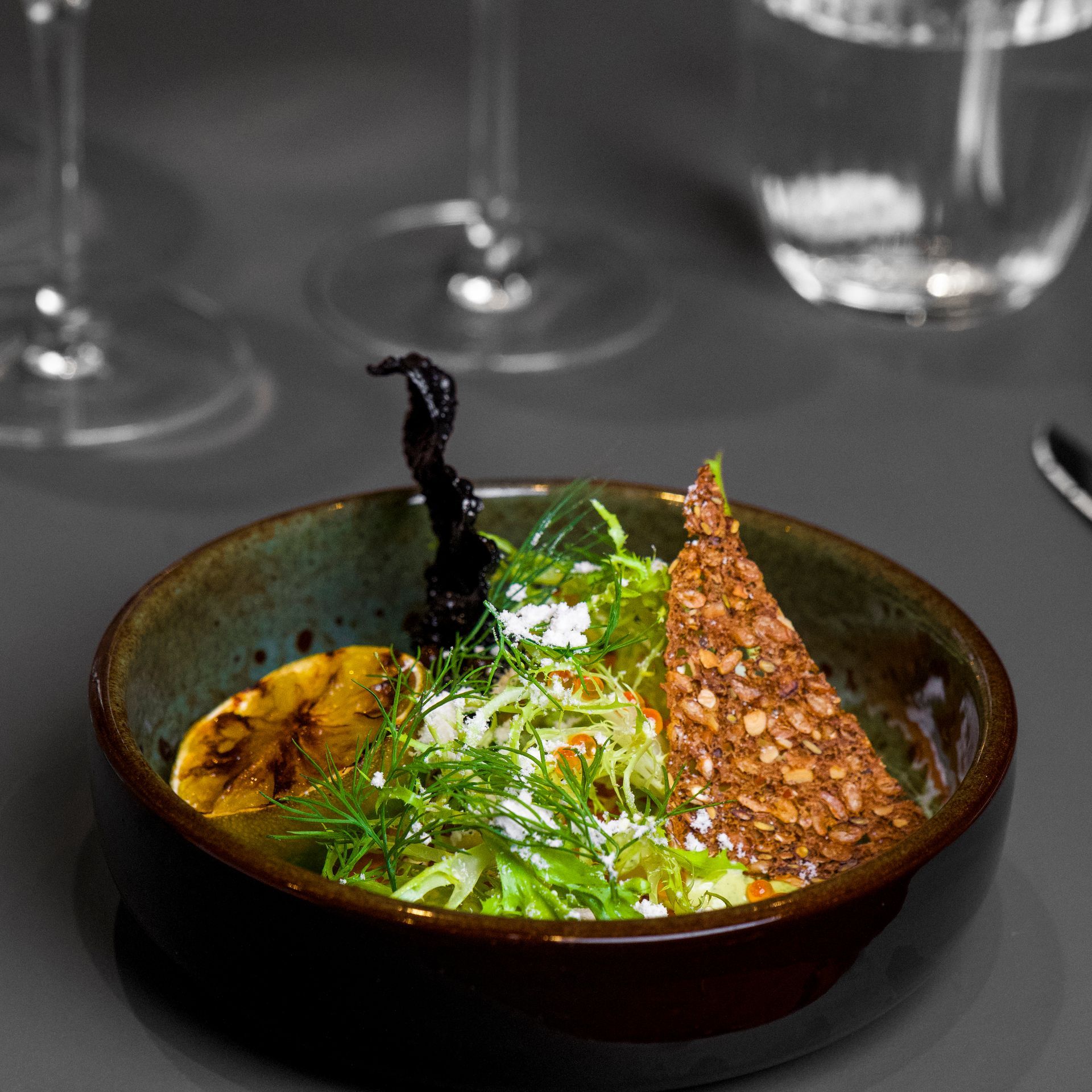 Nærbillede af en lille, rustik skål med en gourmetsalat. Indeholder grillet citron, salatblade, ost og en chips med kerner.