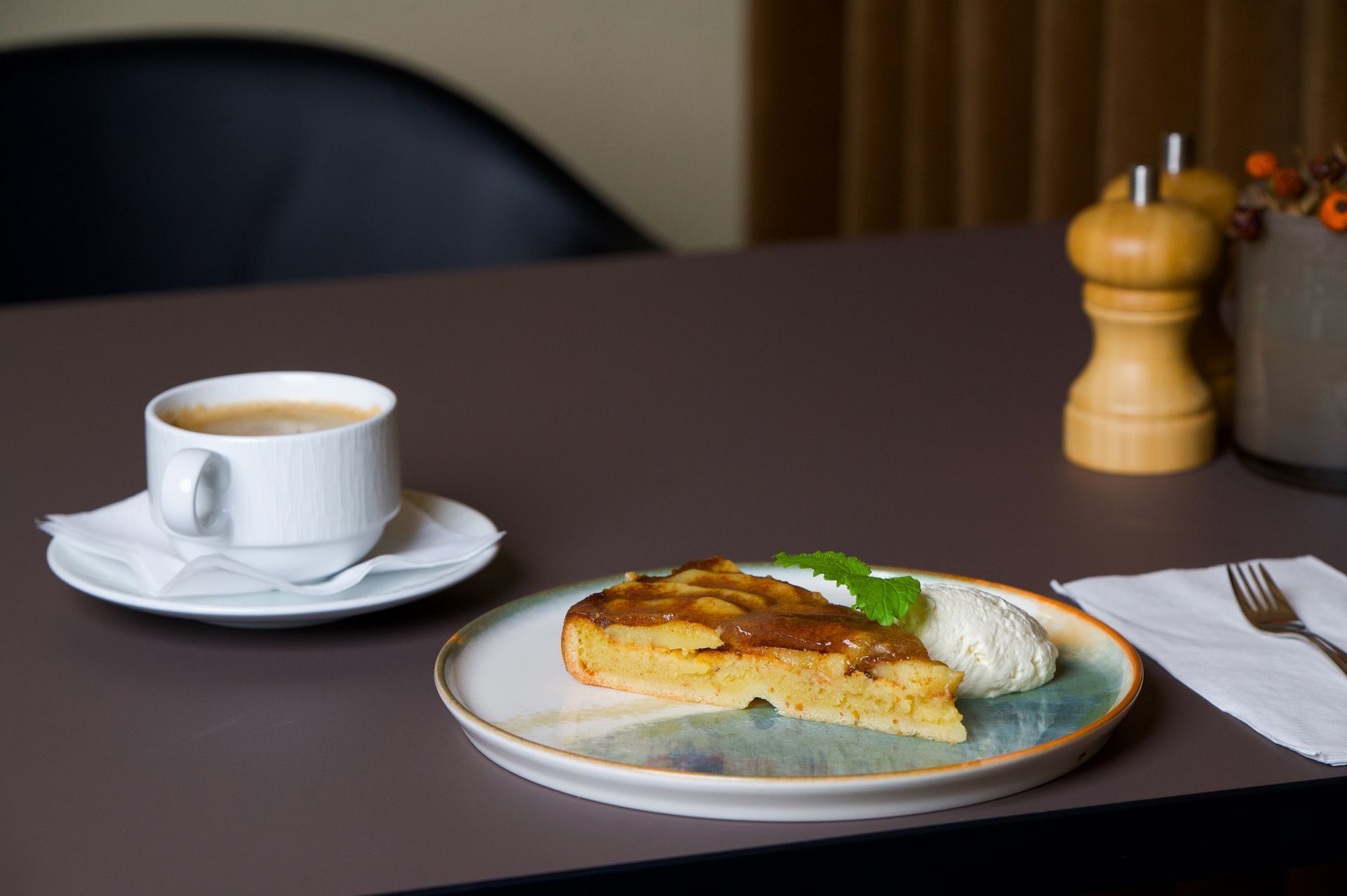 Skive af æblekage med fløde og kaffe på et brunt bord ved siden af ​​en gaffel og peberkværn.