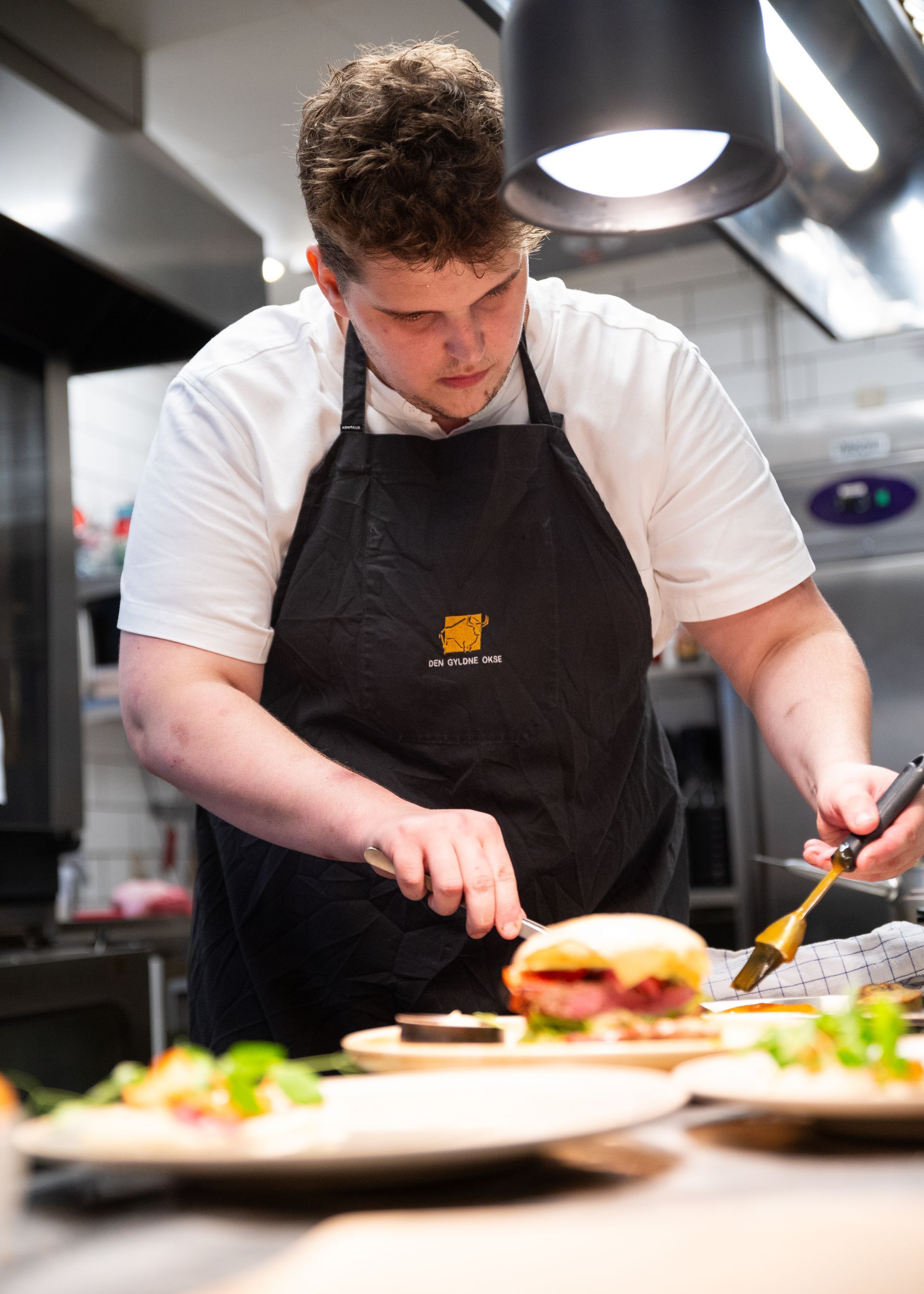Kok i hvid skjorte og sort forklæde skærer en sandwich i et restaurantkøkken.