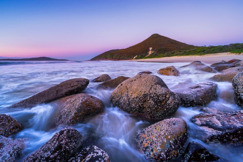 A Rocky Beach with A Mountain in The Background and Waves Crashing on The Rocks  — Concrete Colour Solutions in Port Stephens, NSW