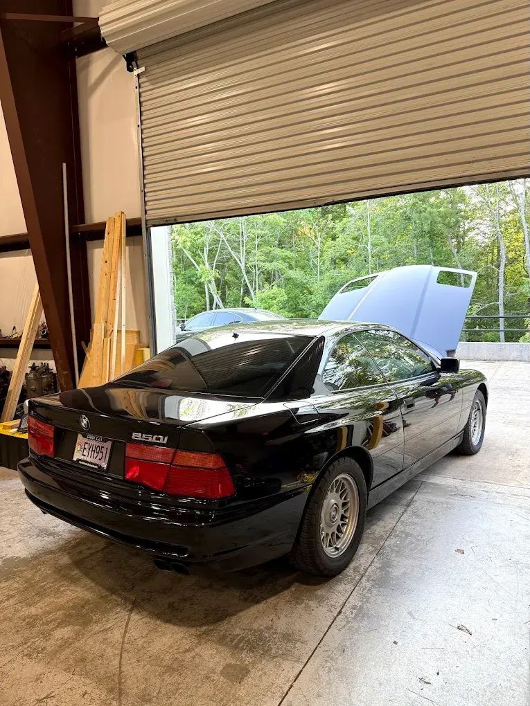 Black BMW coupe with hood up in a garage, facing left, with roll-up door open to trees. | Friendly Automotive