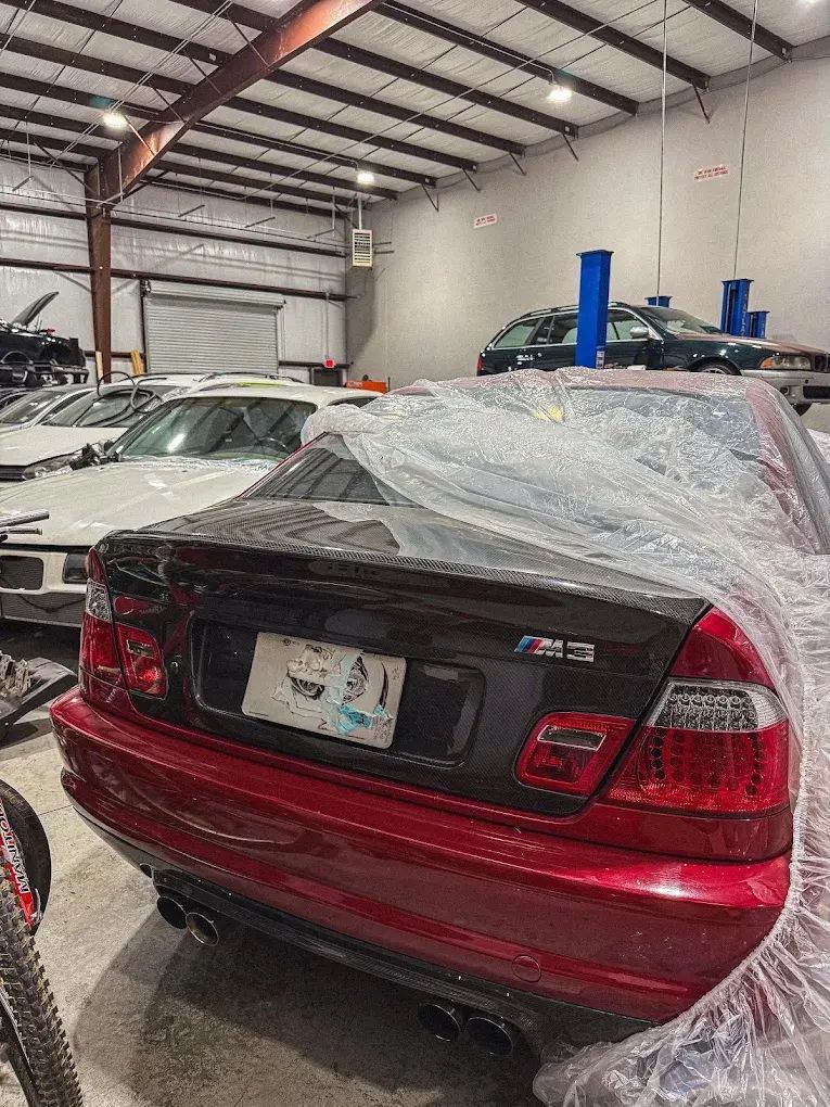 Red BMW M3 sedan under plastic wrap in a warehouse. Other cars are visible in the background. | Friendly Automotive