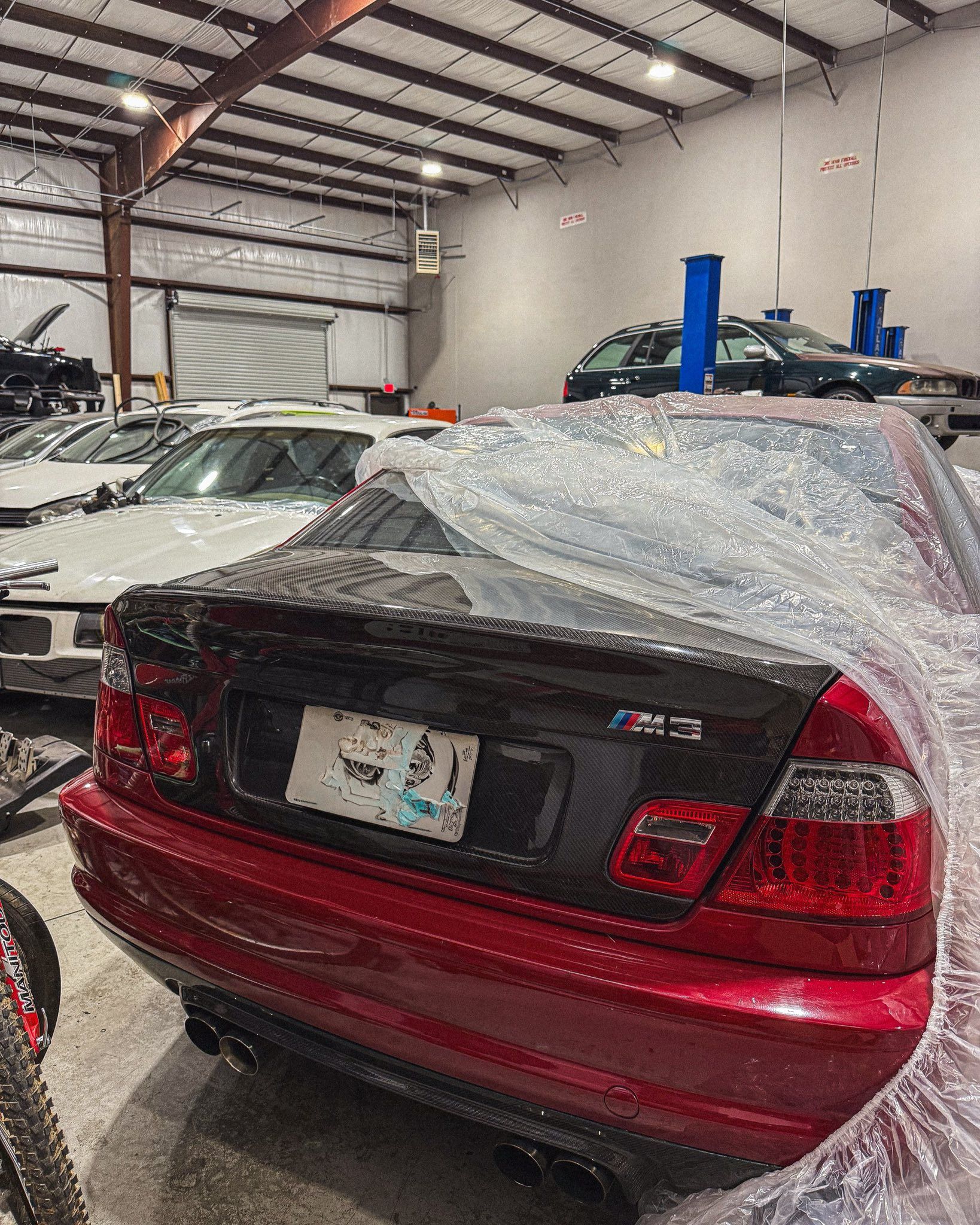 Red BMW M3 under plastic wrap in an auto shop. Other cars and equipment are visible. | Friendly Automotive