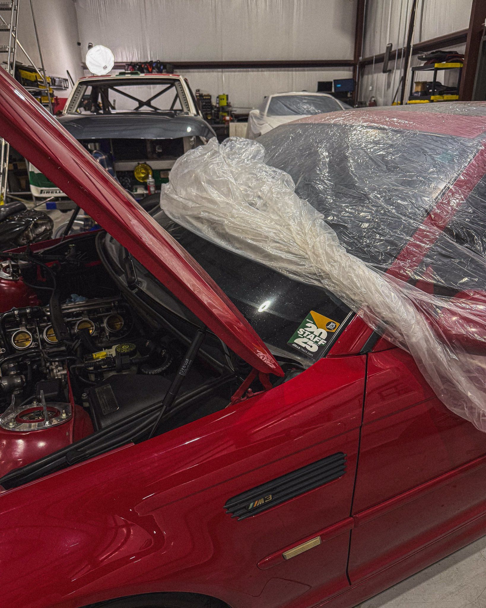 Red car with open hood, covered in plastic wrap, in a garage. Other cars visible in the background. | Friendly Automotive