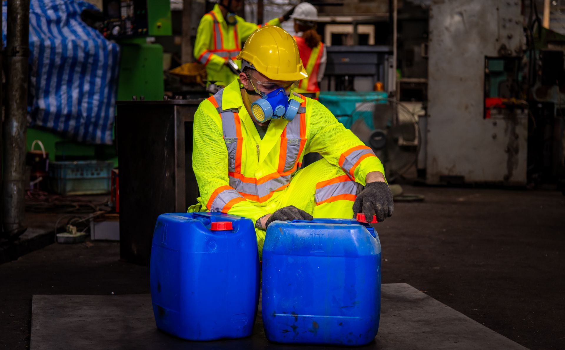 A man in a yellow safety suit is sitting next to two blue barrels.