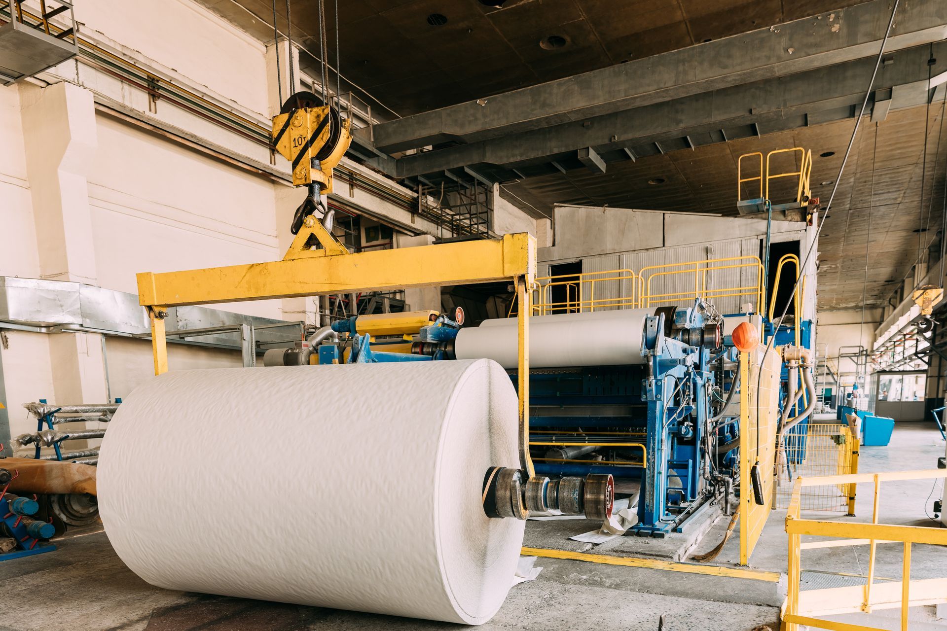 A large roll of paper is being lifted by a crane in a factory.