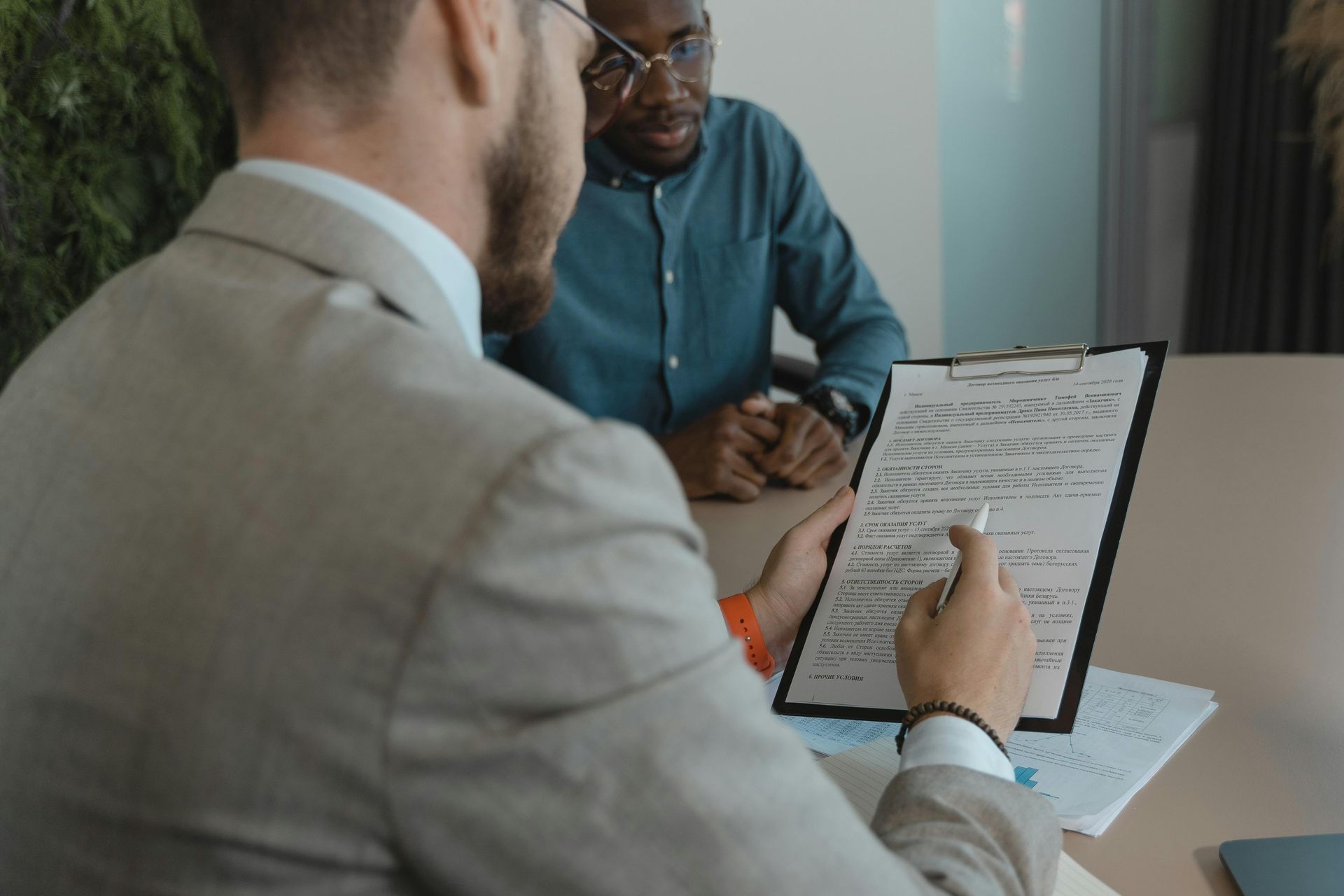 A man is sitting at a table talking to another man while holding a clipboard.