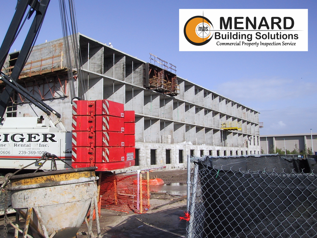 Construction site with a concrete building under construction. Crane, orange safety fencing, and red blocks are visible.